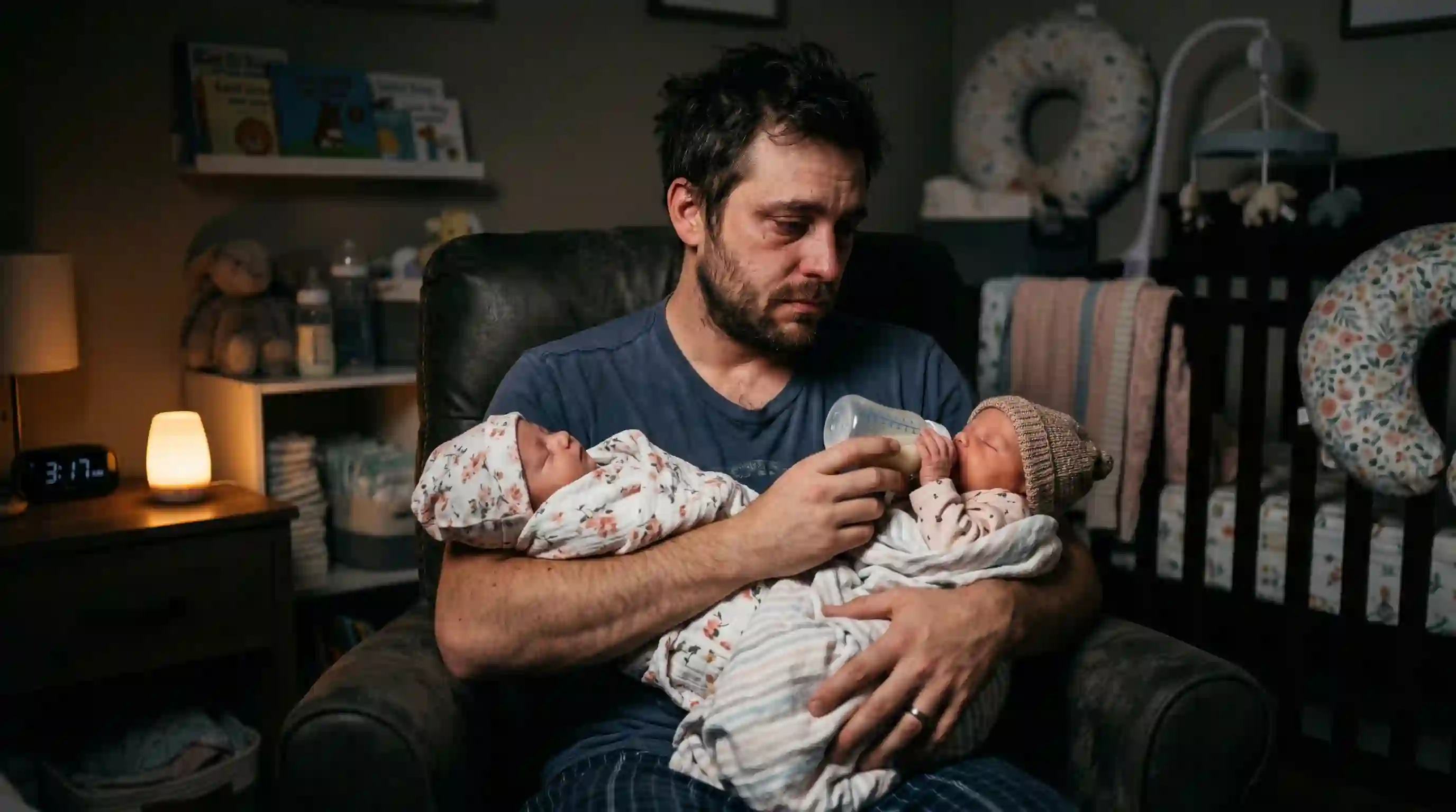 An exhausted young father holding and bottle-feeding his newborn twin daughters in a dimly lit nursery